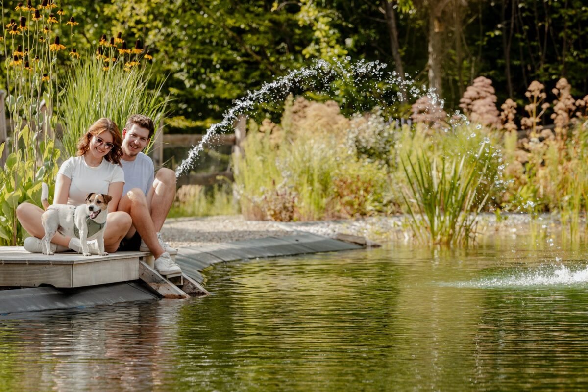 Paar mit Hund sitzt am Steg des Naturteichs, im Wasser eine Fontäne