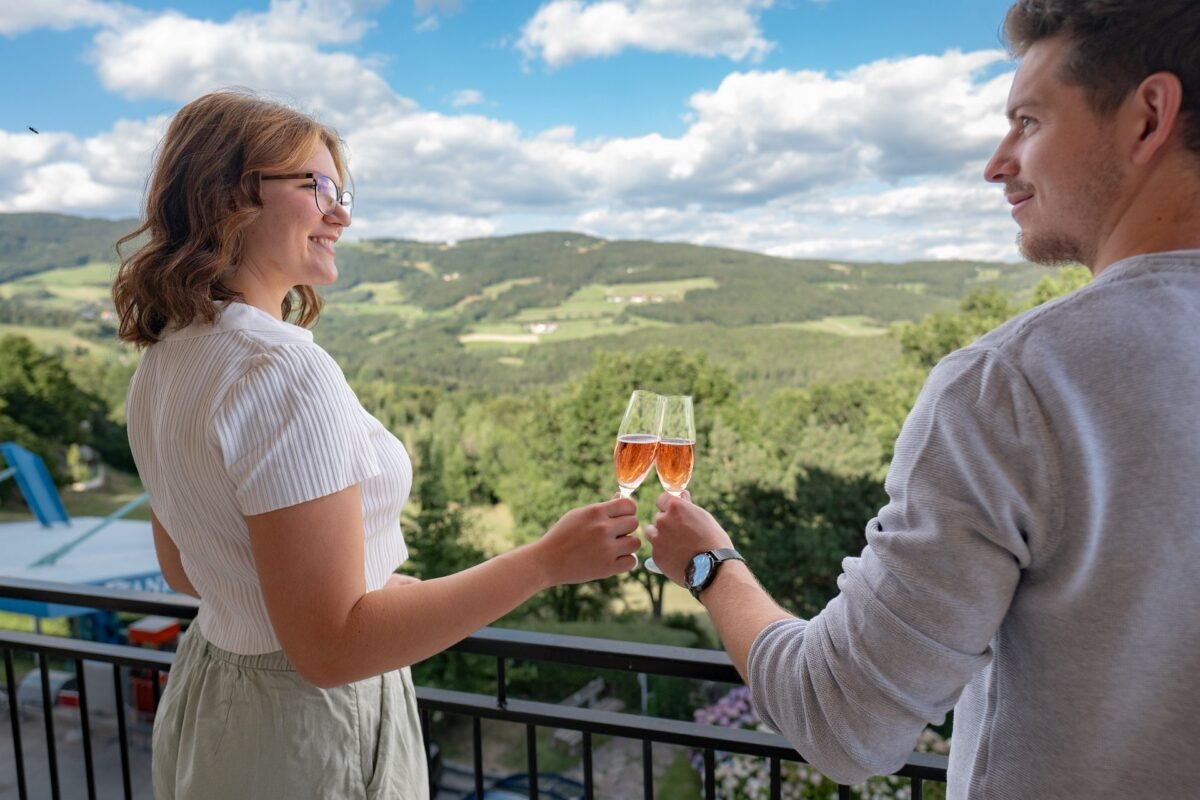 Paar stößt auf dem Balkon mit Sekt an, im Hintergrund die Hügellandschaft